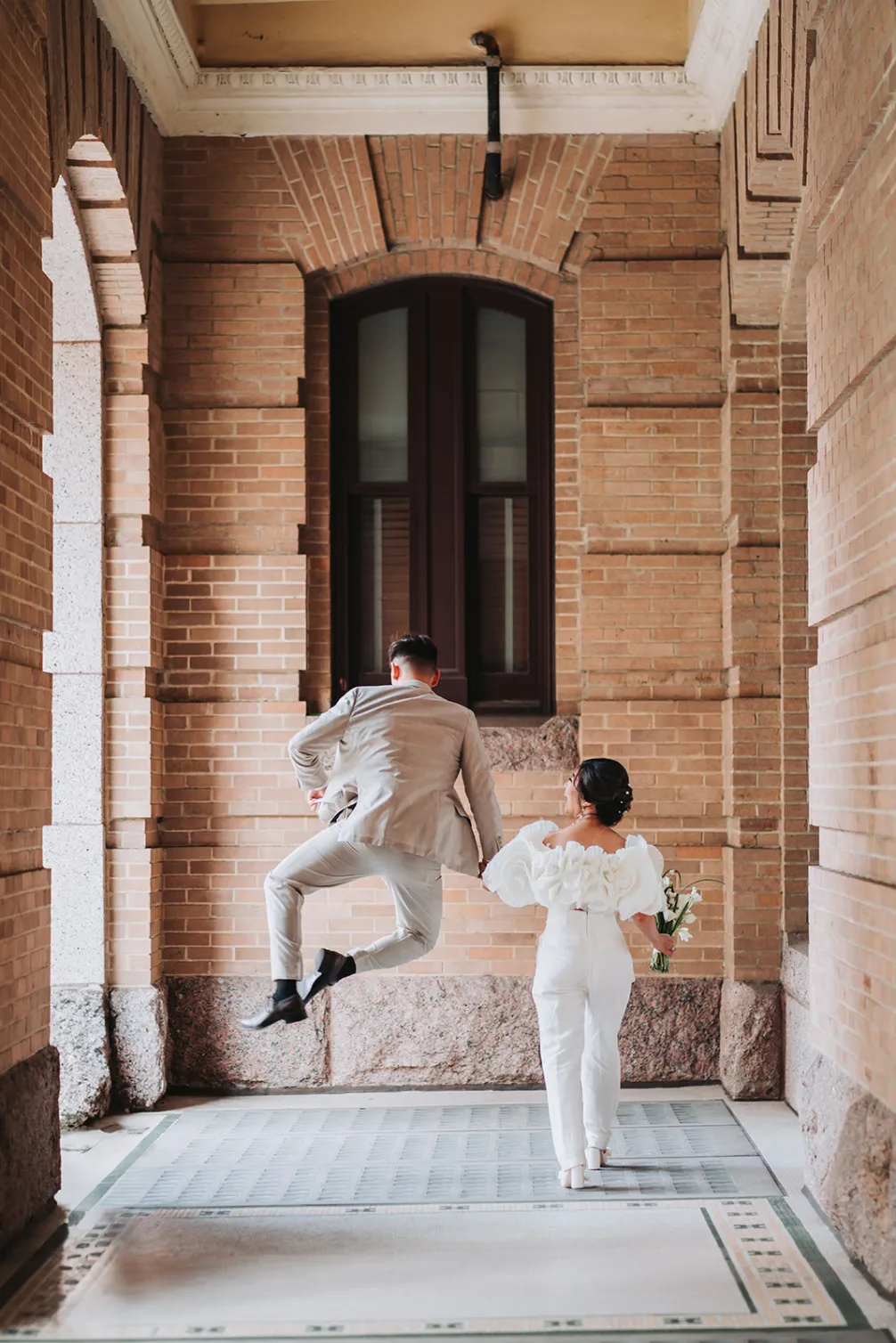 A joyful bride and groom in elegant attire are walking hand in hand along a brick corridor, capturing a lively moment as the groom leaps into the air in celebration.